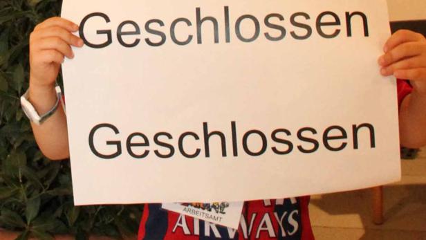 Ein Kind hält ein Schild mit der Aufschrift „Geschlossen“ vor einem Aushang des Arbeitsamtes.
