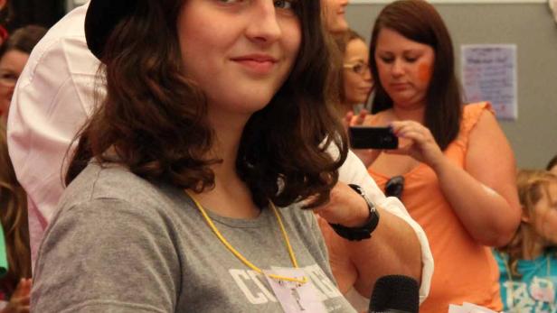 Eine junge Frau mit Hut hält ein Mikrofon und ein Blatt Papier in der Hand.