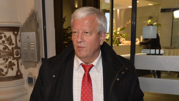 Ein Mann mit grauem Haar und roter Krawatte steht vor einer Tür.