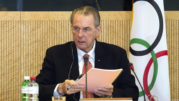 Ein Mann mit Brille sitzt an einem Tisch mit der olympischen Flagge im Hintergrund.