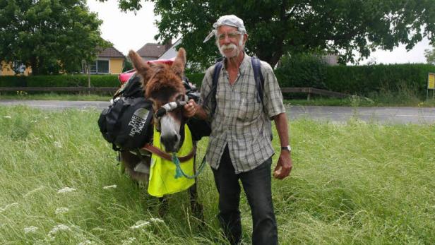 Ein älterer Mann steht mit einem bepackten Esel auf einer Wiese.