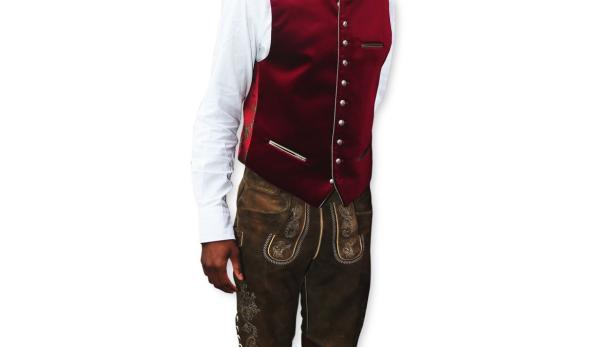 Ein Mann in Tracht mit Lederhose, roter Weste und Hut steht lächelnd da.