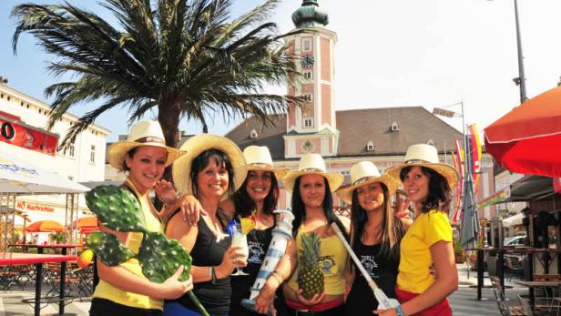 Sechs Frauen mit Strohhüten posieren mit tropischen Requisiten vor einem Gebäude.