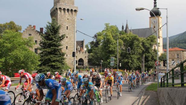 Eine Gruppe Radfahrer fährt auf einer Straße an einer Burg und einer Kirche vorbei.