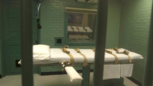 Eine Liege mit Riemen in einem Raum hinter Gitterstäben.