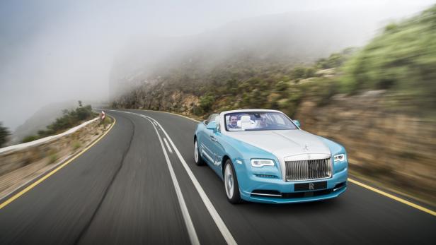 Ein hellblauer Rolls-Royce fährt auf einer kurvenreichen Straße entlang einer Küste.