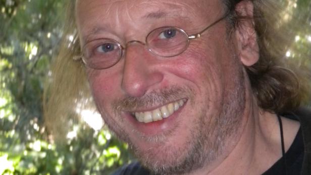 Ein lächelnder Mann mit Brille und blonden Haaren vor grünem Hintergrund.