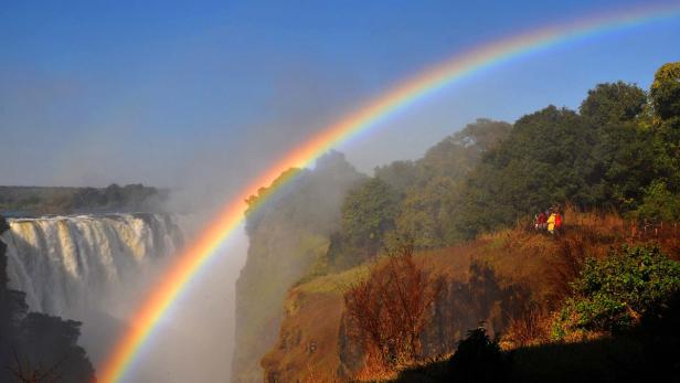 Ein Regenbogen überspannt einen Wasserfall und eine bewaldete Klippe.