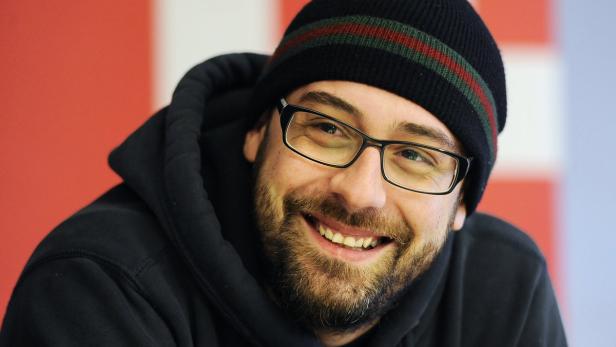 Ein lächelnder Mann mit Brille und Mütze vor einem rot-weißen Hintergrund.