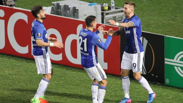 Drei Fußballspieler in blauen Trikots jubeln auf dem Spielfeld.
