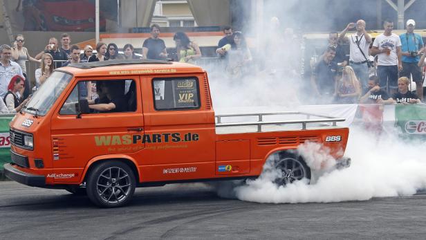 Ein orangefarbener VW-Bus macht einen Burnout vor einer Zuschauermenge.
