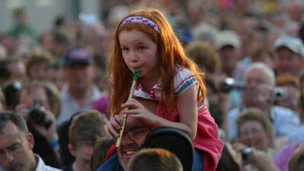 Ein Mädchen mit roten Haaren spielt auf einer Tin Whistle, getragen auf den Schultern eines Mannes.