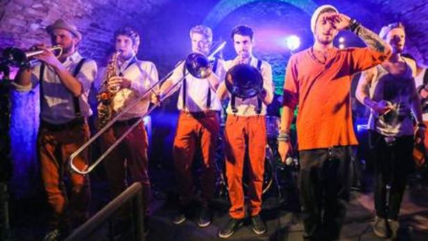 Eine Band spielt in einer dunklen Umgebung mit Trompete, Saxophon, Posaune und Schlagzeug.