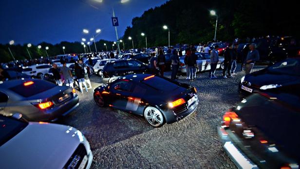 Ein dunkler Audi R8 steht auf einem Parkplatz voller Autos und Menschen.