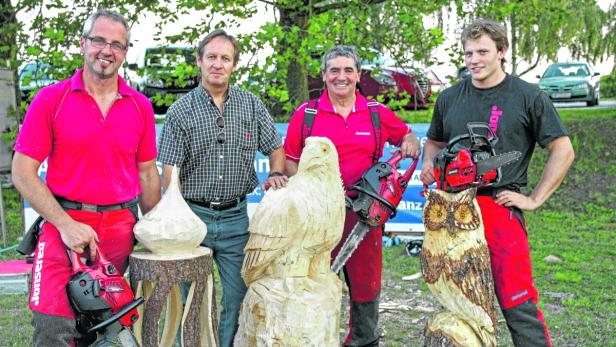 Vier Männer posieren mit Kettensägen und geschnitzten Holzfiguren, darunter ein Adler und eine Eule.