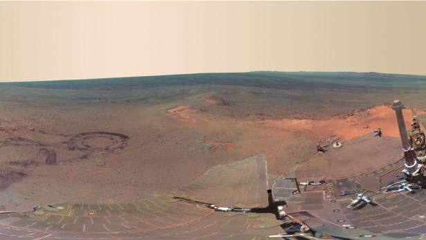 Panoramaaufnahme der Marsoberfläche mit Spuren im Sand und Teilen eines Rovers im Vordergrund.