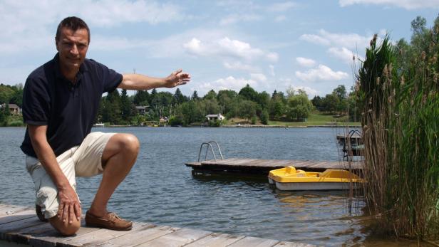 Ein Mann kniet auf einem Holzsteg an einem See und deutet mit dem Arm in die Ferne.
