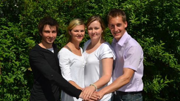 Vier junge Leute stehen vor einem grünen Busch und halten ihre Hände zusammen.
