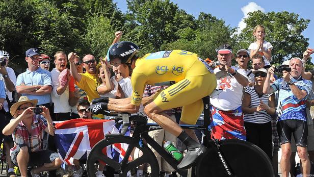Ein Radfahrer im gelben Trikot fährt ein Rennen, angefeuert von Zuschauern mit Union-Jack-Flaggen.