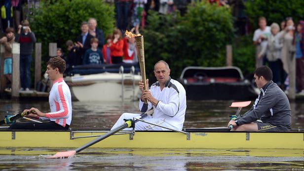 Drei Ruderer in einem gelben Boot, der mittlere hält die olympische Fackel.