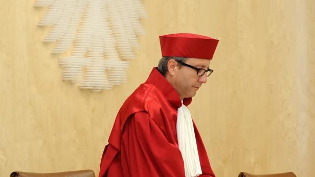Ein Richter in roter Robe und mit Brille steht vor einer hellen Holzwand.