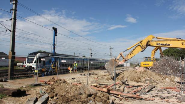 Ein Bagger steht neben einer Baustelle an einem Bahnhof, wo ein Zug hält.