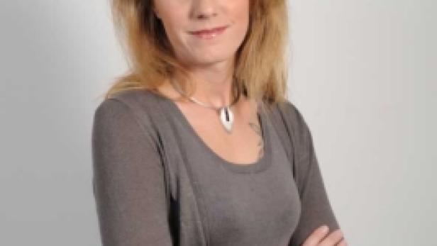 Eine Frau mit blonden Haaren und verschränkten Armen vor einem hellen Hintergrund.