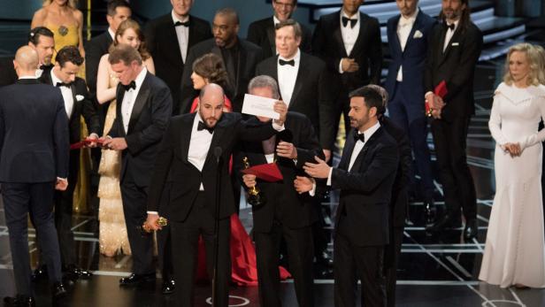 Ein Mann im Anzug auf der Oscar-Verleihung hält eine Karte hoch.