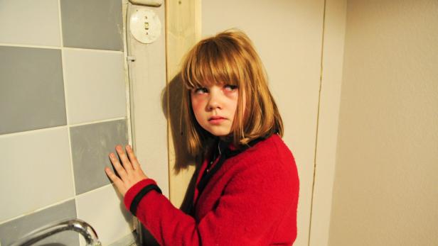 Ein junges Mädchen in roter Jacke steht an einer gefliesten Wand.