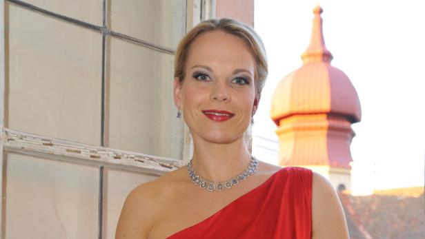 Eine Frau in rotem Kleid und Schmuck vor einem Fenster mit Blick auf ein Gebäude.