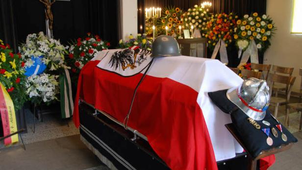 Ein Sarg, bedeckt mit der österreichischen Flagge, steht in einer Kirche, geschmückt mit Blumen und einem Helm.