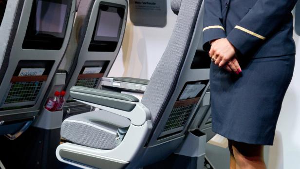 Eine Flugbegleiterin steht neben einem Flugzeugsitz mit eingebautem Bildschirm und Magazinhalter.