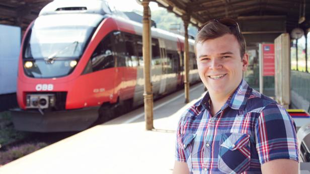 Ein lächelnder Mann steht an einem Bahnsteig vor einem roten ÖBB-Zug.