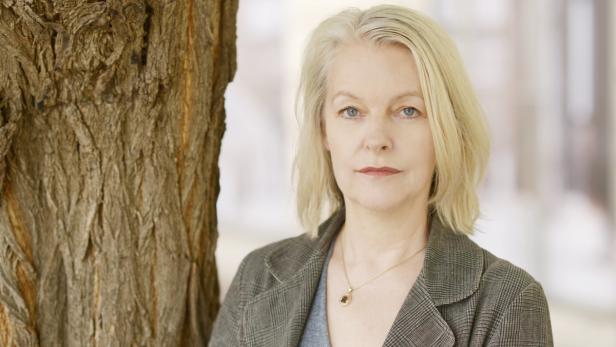Eine Frau mit blonden Haaren und einem grauen Blazer steht neben einem Baumstamm.