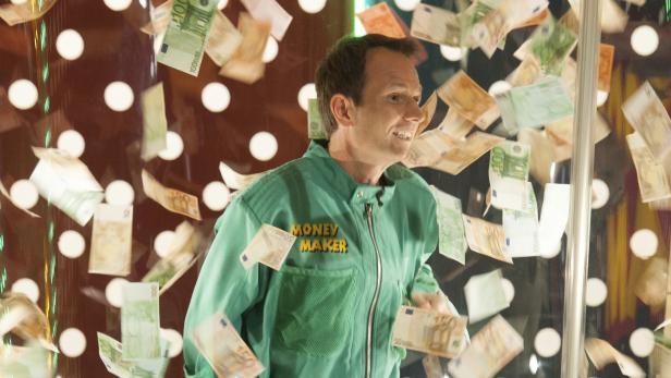 Ein Mann in einer „Money Maker“-Jacke steht in einer Kabine, in der Geldscheine umherwirbeln.