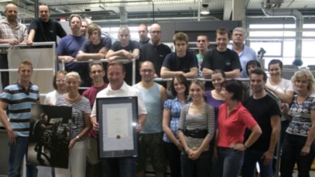 Eine Gruppe von Menschen posiert mit einem gerahmten Dokument und einem Poster in einer Fabrikhalle.