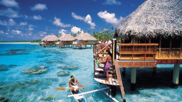 Ein Mann fährt mit einem Kanu an strohgedeckten Bungalows auf dem Wasser vorbei.