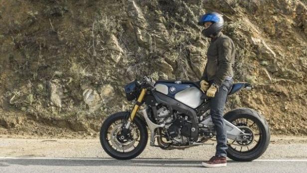 Ein Mann mit Helm steht neben einem Motorrad vor einer Felswand.
