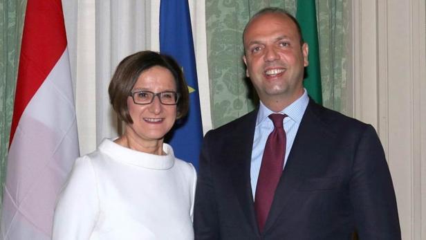 Eine Frau in Weiß und ein Mann im Anzug posieren vor Flaggen.