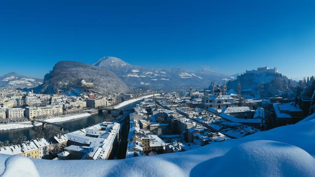 Winterliche Panoramaansicht von Salzburg mit schneebedeckten Dächern und Bergen.