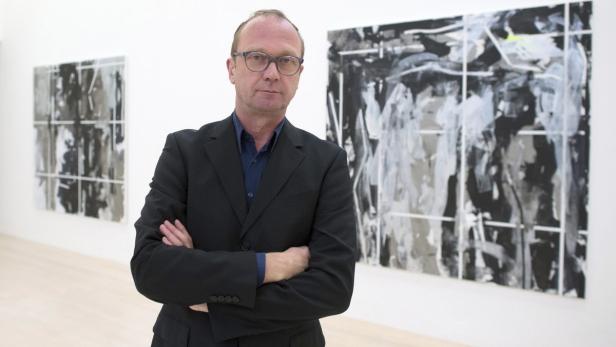 Ein Mann mit Brille und dunklem Anzug steht vor zwei abstrakten Gemälden.