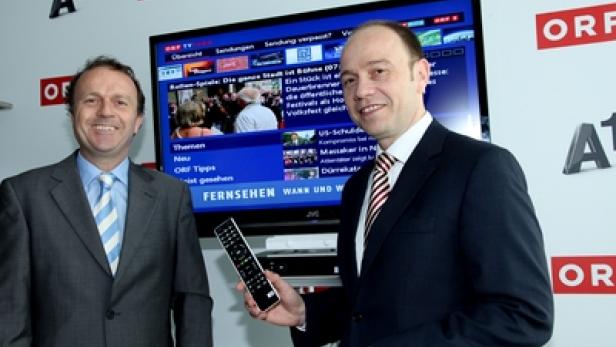 Zwei Männer stehen vor einem Fernseher mit dem ORF-Logo und halten eine Fernbedienung.