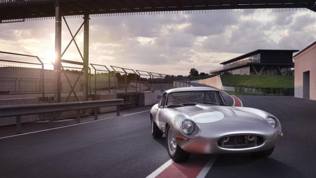 Ein silberner Jaguar E-Type auf einer Rennstrecke unter einer Fußgängerbrücke.