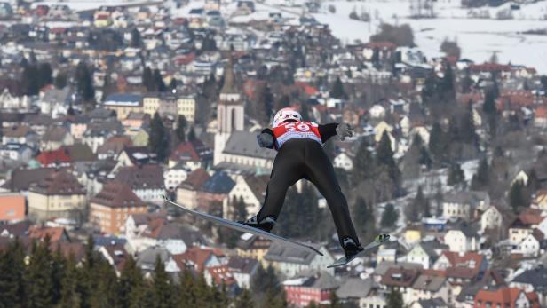 Ein Skispringer in der Luft vor einer Stadtkulisse.