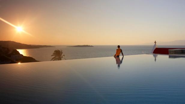 Ein Mann sitzt am Rand eines Infinity-Pools und blickt auf das Meer bei Sonnenuntergang.