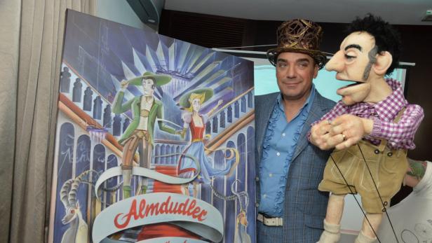 Ein Mann mit Zylinder steht neben einer Almdudler-Werbung und einer Marionette.