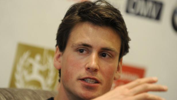 Ein Mann mit braunen Haaren spricht vor einer Wand mit Logos.