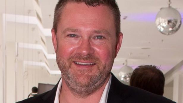 Porträt eines lächelnden Mannes mit Bart in einem dunklen Anzug.