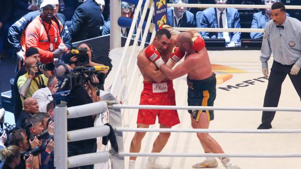Zwei Boxer kämpfen im Ring, umringt von Fotografen und Zuschauern.