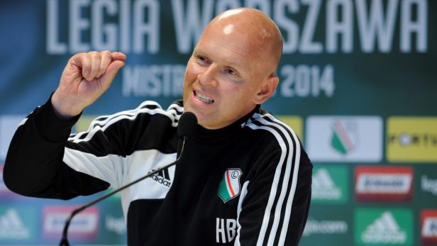 Ein Mann in Sportkleidung gestikuliert vor einem Legia Warschau-Banner.